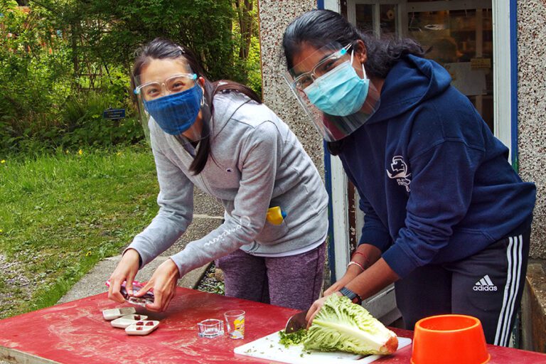 dos chicas cortando comida en una mesa, situada en el exterior de un edificio, como parte de su voluntariado en el extranjero