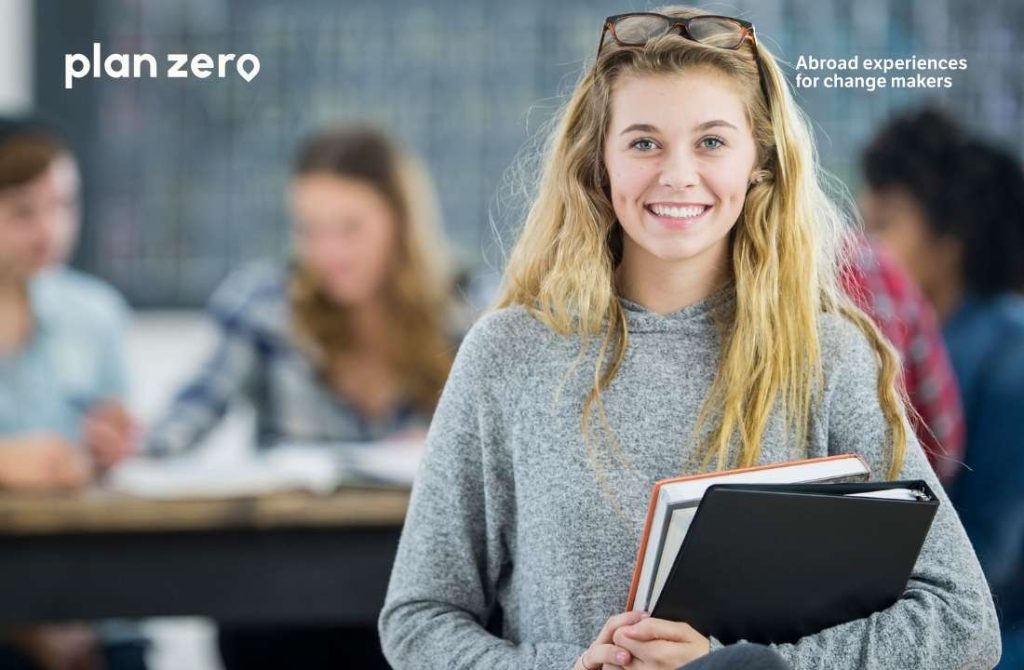 Estudiante joven y rubia, sujetando varias carpetas, mirando a cámara. Al fondo, desenfocado, se ven otros compañeros en el aula.