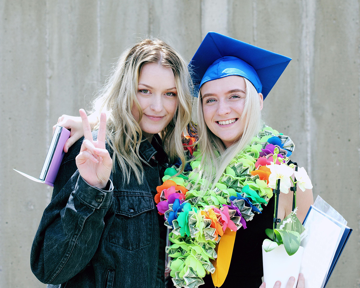 dos jóvenes abrazadas y sonrientes, una de ella lleva un birrete en la cabeza, guirnaldas en el cuello y una flor en la mano