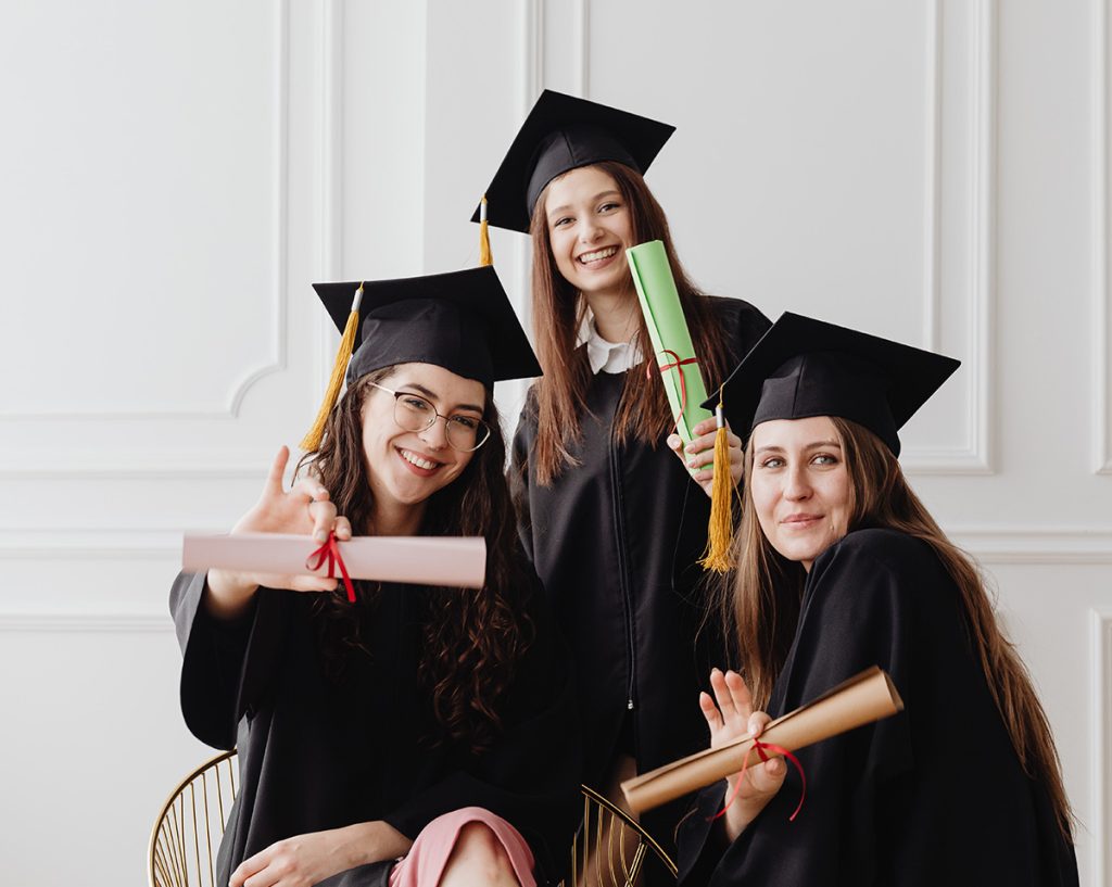 tres chicas jóvenes vestidas con toga y birrete, sonrientes, mostrando sus diplomas