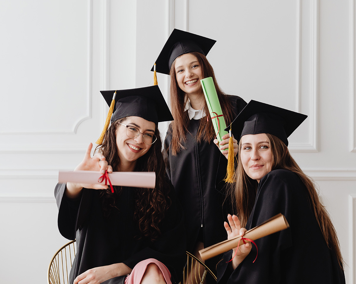 tres chicas jóvenes vestidas con toga y birrete, sonrientes, mostrando sus diplomas